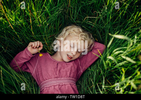 Smiling sleeping dreaming Caucasian girl with closed eyes lying in grass holding yellow bulbous buttercup flower. Happy child kid enjoying summer. Vie Stock Photo