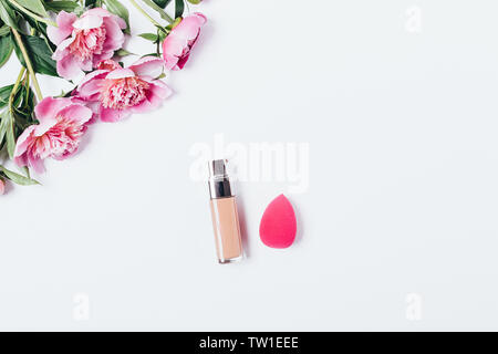 Makeup blending sponge applicator and facial foundation cream near bouquet of pink peonies on white background, flat lay. Stock Photo