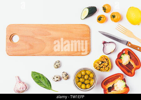 Food background of wooden board next to tomatoes, cucumbers, bell peppers, nuts, greens and quail eggs, flat lay composition on white kitchen table. Stock Photo