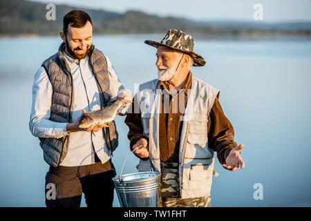 Happy grandfather with adult sun standing together with fresh caught fish on the pier near the lake early in the morning Stock Photo