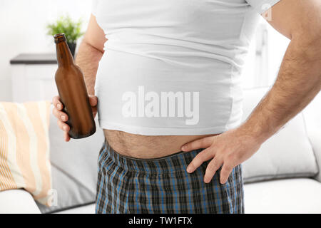 Man with big belly holding bottle of beer at home Stock Photo