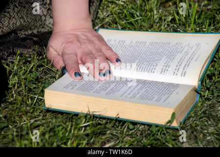 Nice fashion woman lies on green grass and reading an interesting book in city park at summer day. Handsome student woman is sitting and reads book. Stock Photo