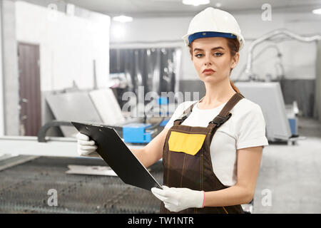 Front view of serious young female engineer looking at camera and posing while working on metal factory. Beautiful woman keeping folder in hands and checking computer. Concept of industry. Stock Photo
