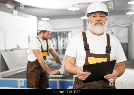 Front view of professional male wearing uniform and helmet keeping folder, looking at camera and posing while working in metal industry. Engineer controlling processes of devices. Concept of factory. Stock Photo