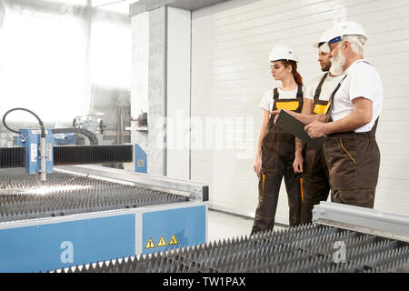 Side view of three engineers standing near cnc, talking and discussing work process on metal factory. Male and female mechanics in uniforms and helmets looking at plasma cutting. Concept of industry. Stock Photo