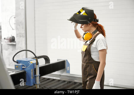 Side view of young female mechanic in protective mask and uniform standing near cnc and controlling process of plasma cutting on metal factory. Concept of technology and machinery. Stock Photo