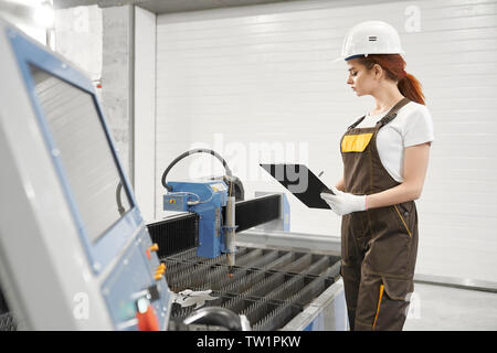 View from side of attractive female engineer in process of controlling plasma cutting device. Young woman in helmet and uniform standing near equipment and writing data. Concept of metalwork. Stock Photo