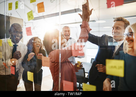 Two diverse businesspeople high fiving together after a brainstorming session in an office with laughing colleagues looking on Stock Photo