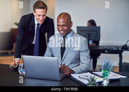 Two smiling diverse businessmen using a laptop together while working at a desk in a modern office Stock Photo