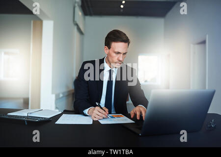 Focused businessman going over paperwork and working on a laptop while sitting at his desk in a bright modern office Stock Photo