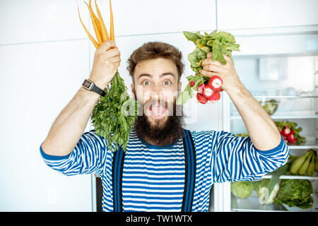 Portrait of a well-looking man with carrot and radish in front of the fridge full of fresh vegetables and fruits at home Stock Photo