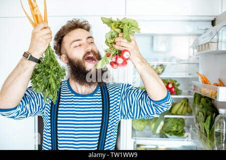 Portrait of a well-looking man with carrot and radish in front of the fridge full of fresh vegetables and fruits at home Stock Photo