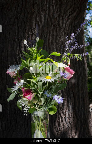 Beautiful Rustic Wedding Floral Arrangement of Pink Roses, Poppies and Daisies against a background of Bark of an Oak Tree in bright sunshine Stock Photo