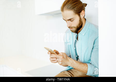 Handsome man using phone while sitting on the white kitchen at home Stock Photo