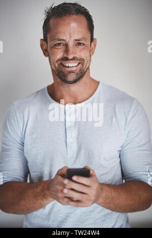 Casually dressed male entrepreneur reading a text message on his cellphone and smiling while standing against a gray background Stock Photo