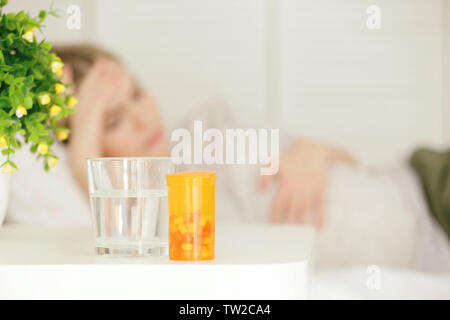 Glass of water, box with pills on table and blurred woman suffering from headache on background Stock Photo