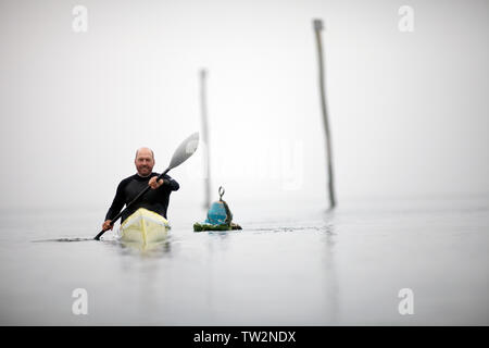 Male kayaker smiles for a portrait as he paddles past tall mooring poles and a buoy in a foggy harbour. Stock Photo