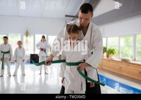 Trainer putting belt. Dark-haired aikido trainer putting belt on waist of cute blonde girl Stock Photo