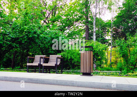 Two individual wooden park chairs and  trashcan on sidewalk edge in green well-kept park Stock Photo