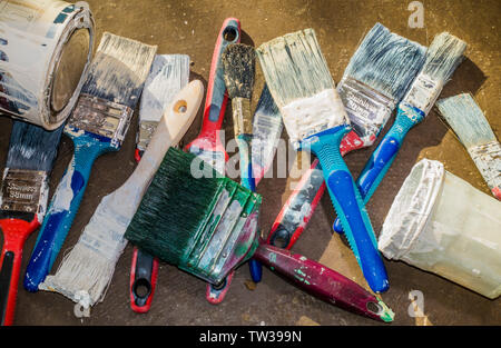 A pile of old, used paintbrushes with hard bristles and coated paint, ready to be thrown away, along with an old glass jar and a tin of paint. Stock Photo