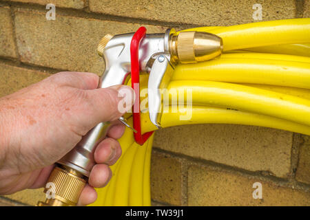 A man’s hand replacing the spray gun attached to the end of a yellow coiled garden hose, which is hanging over hooks attached to a wall. Stock Photo