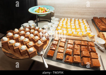 Small assorted cakes lined up decorated on dessert buffet along with fruit salad. Sweet paradise. Stock Photo