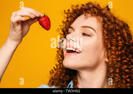 curly red-haired woman with open mouth wants to eat delicious strawberries on a yellow background. Stock Photo