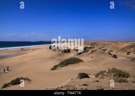 View over sand dunes with green ocean on (Playa del Aljibe) on white village on steep cliff (El Cotillo) - North Fuerteventura Stock Photo