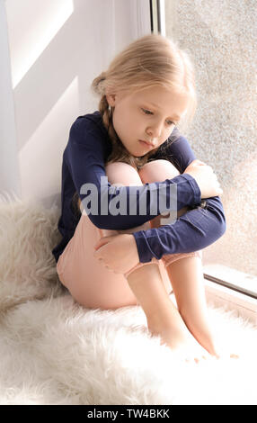 Cute sad girl sitting on window sill at home Stock Photo