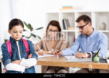 selective focus of upset kid with backpack standing with crossed arms near parents in office Stock Photo