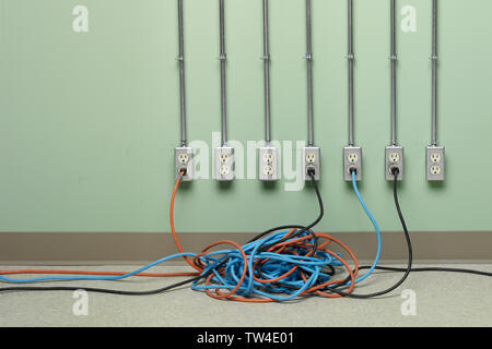 Blue, orange and black tangled extension cords plugged into row of AC electrical outlets on green wall. Stock Photo