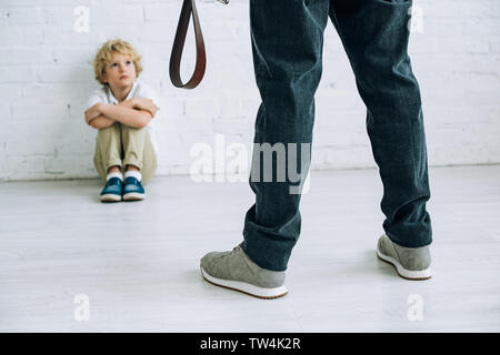 partial view of abusive father holding belt and son sitting on floor Stock Photo