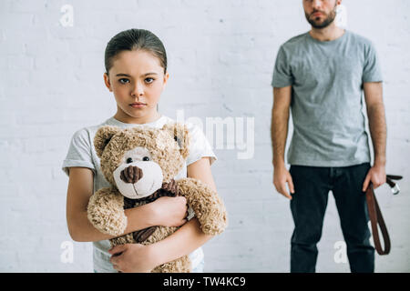 partial view of abusive father with belt and sad daughter holding teddy bear Stock Photo