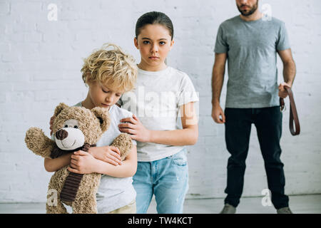 partial view of abusive father with belt and sad kids with teddy bear Stock Photo