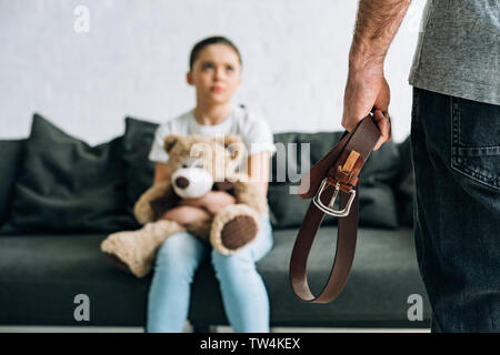 partial view of abusive father holding belt and scared daughter with teddy bear sitting on sofa Stock Photo