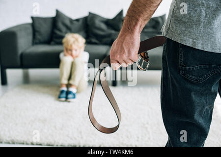 partial view of abusive father holding belt and sad son sitting on carpet Stock Photo