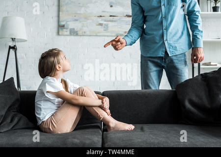 cropped view of man pointing with finger at upset kid sitting on sofa at home Stock Photo