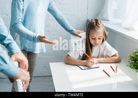 cropped view of parents standing near upset daughter holding pencil at home Stock Photo