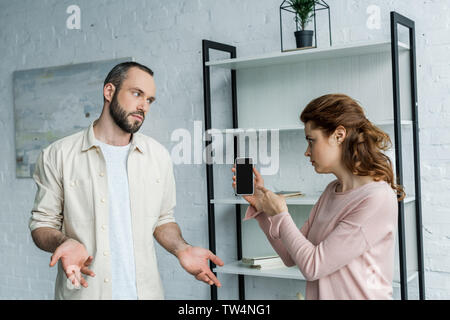 offended woman pointing with finger at smartphone with blank screen near man at home Stock Photo