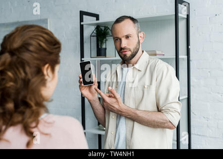 selective focus of offended man pointing with finger at smartphone with blank screen near woman Stock Photo