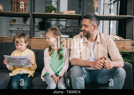 cute boy reading sport news newspaper while sitting on sofa near sister and dad Stock Photo