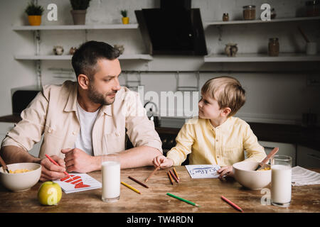 father and son drawing while sitting at kitchen table with served breakfast Stock Photo