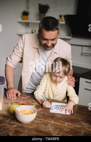 happy father hugging adorable son sitting at kitchen table and drawing fathers day greeting card Stock Photo