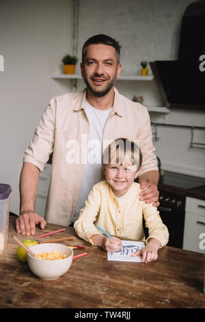 happy father hugging adorable son sitting at kitchen table and drawing fathers day greeting card Stock Photo