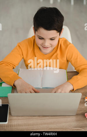 smiling focused boy using laptop while sitting at desk with copy book and doing schoolwork at home Stock Photo