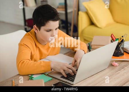 attentive boy using laptop while sitting at wooden desk and doing homework Stock Photo