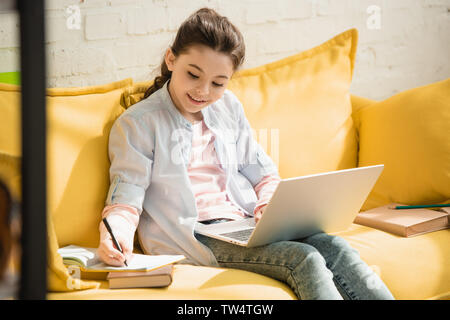 smiling attentive kid writing in copy book and using laptop while sitting on sofa at home Stock Photo