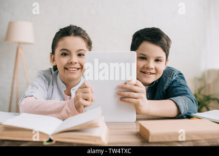 cheerful children holding digital tablet and looking at camera while sitting at table with books together Stock Photo