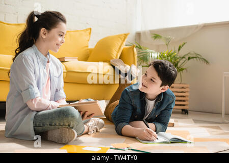 smiling boy lying on floor and writing in copy book near sister sitting with book Stock Photo