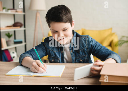 smiling boy writing in notebook and using smartphone while doing schoolwork at home Stock Photo